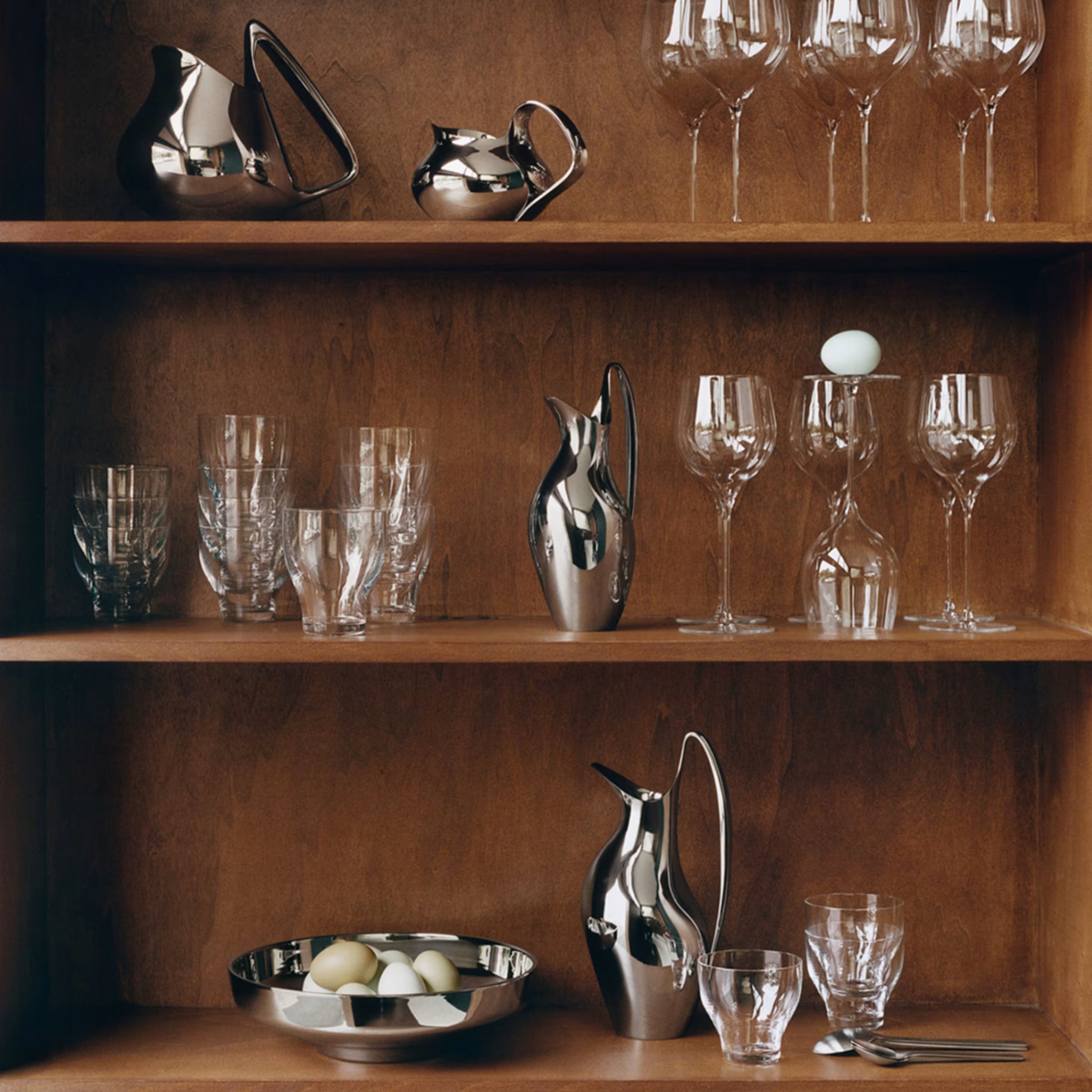 Una jarra Koppel de 1.2 L de Georg Jensen exhibida de manera sofisticada en una estantería de madera junto a cristalería fina.