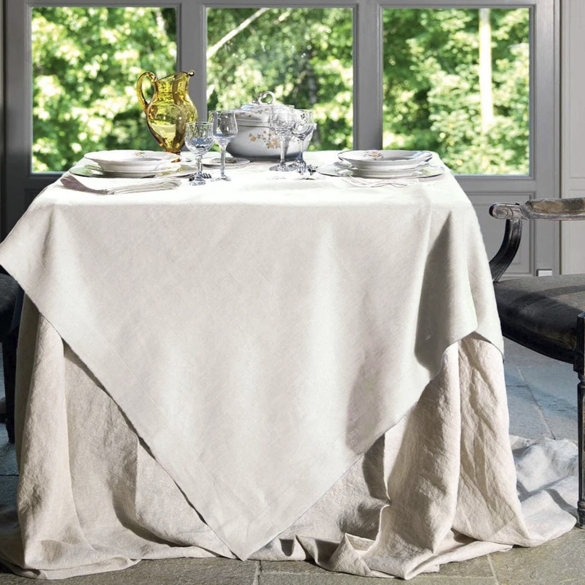Mantel Tiziano Crema de Tessitura Toscana Telerie vistiendo una mesa de comedor elegante con vajilla de porcelana, copas de cristal y cubiertos dorados.