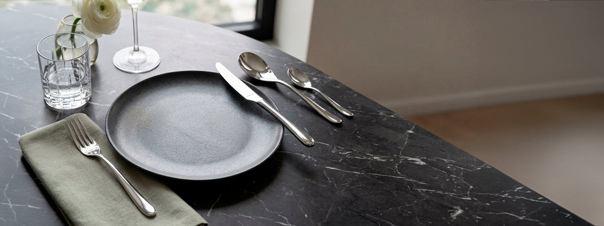 Juego de cubiertos de acero inoxidable pulido Amefa línea Newton junto a plato cerámico sobre mesa de mármol negro