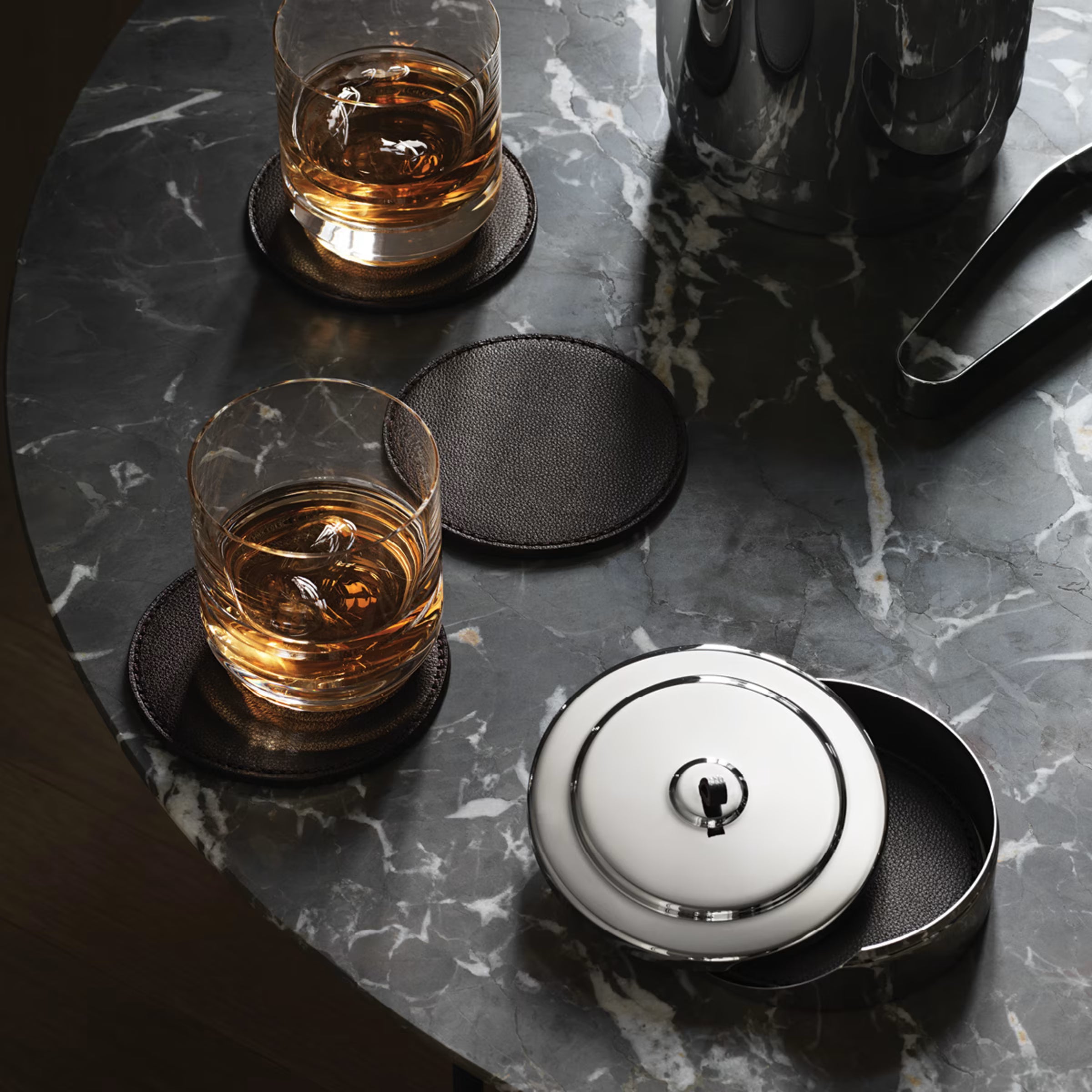 uesta en escena del set Manhattan de Georg Jensen sobre una mesa de mármol oscuro, con dos portavasos de cuero sirviendo de base para vasos de whisky cortos.